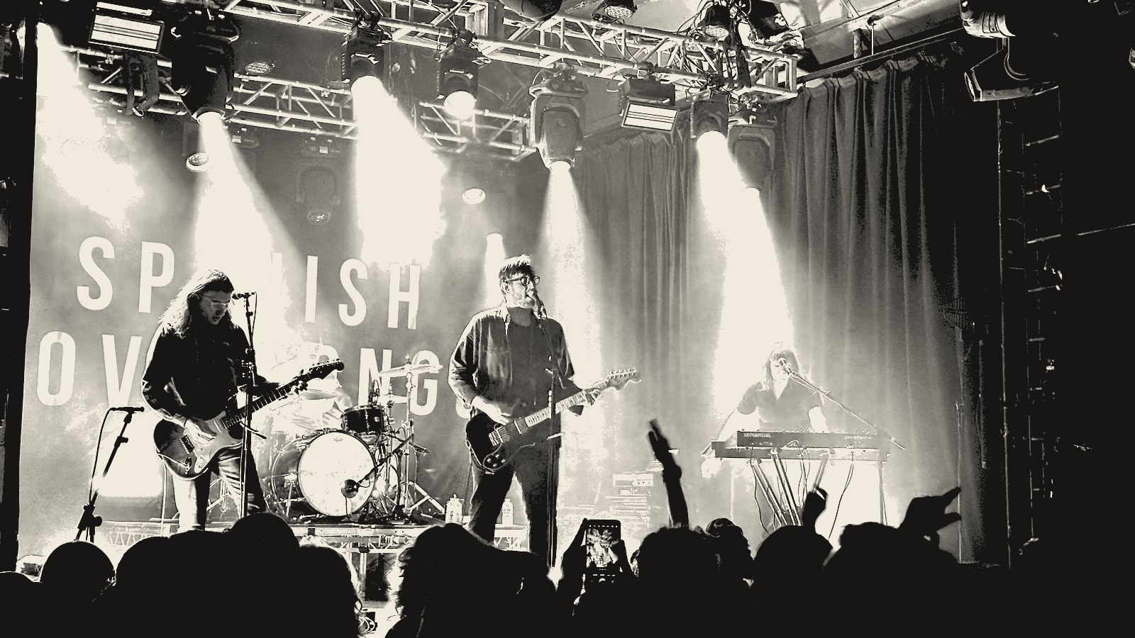 Spanish Love Songs on stage, the band's name printed large on the backdrop behind them, the crowd's hands up in the foreground